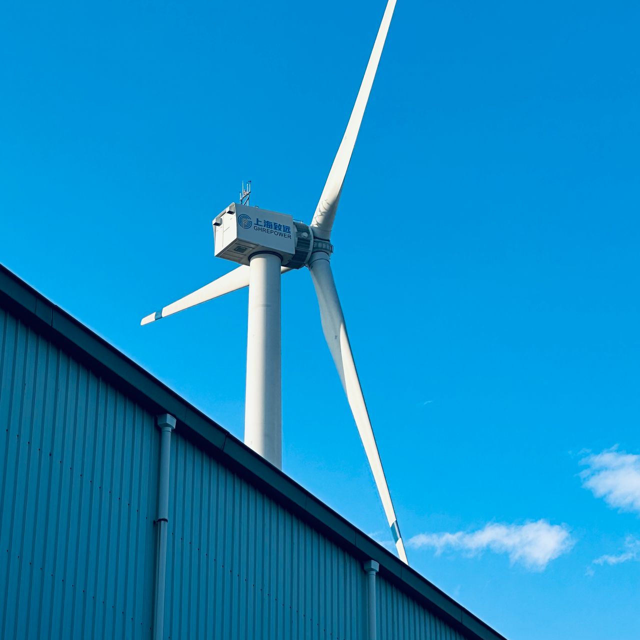 A wind turbine beside a building under a bright blue sky, highlighting renewable energy technology.