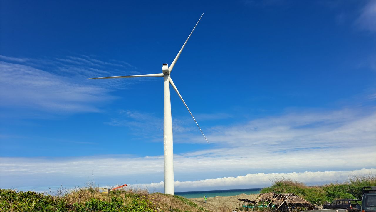 A wind turbine stands tall under a bright blue sky near the ocean, representing renewable energy.