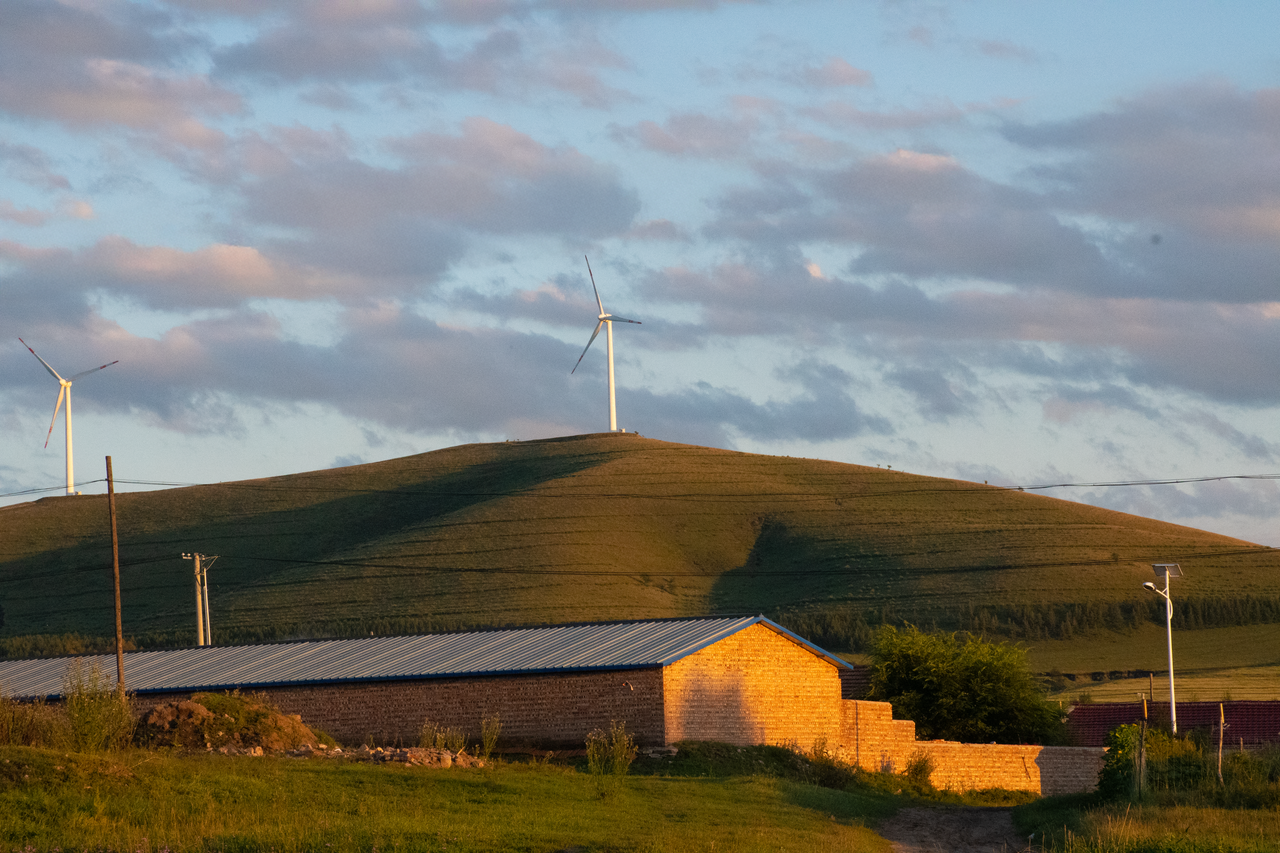 Wind turbines and house on grassy hill during sunset, showcasing renewable energy and rural landscape.
